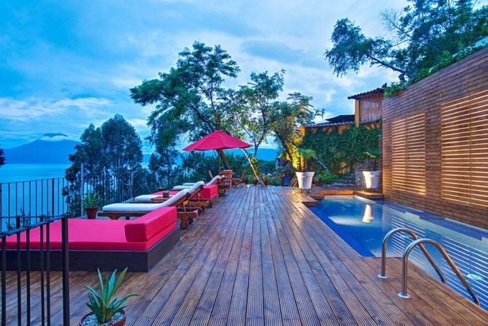 A wooden deck hosts red lounge chairs beside a pool, surrounded by trees, overlooking a scenic view of water and distant mountains.
