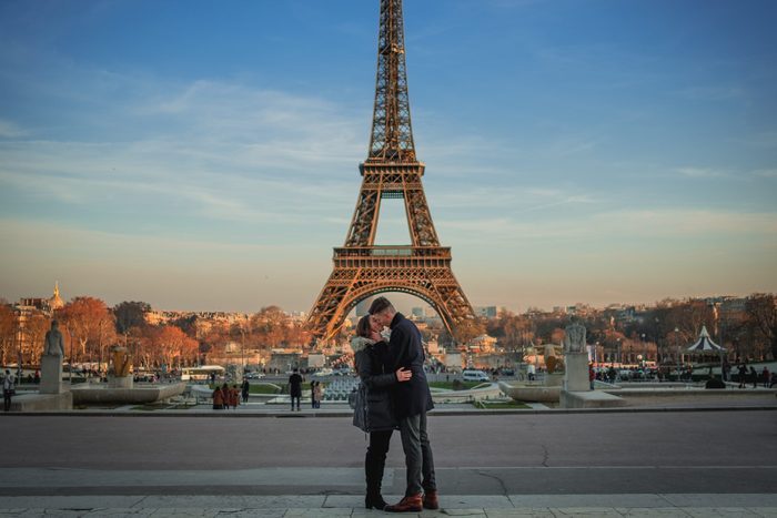 A couple embraces in front of the Eiffel Tower, surrounded by a park with fountains and autumn-colored trees under a clear blue sky.