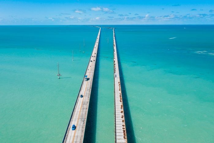 A long bridge stretches across turquoise water, with cars driving over it under a clear blue sky, surrounded by vast ocean views.