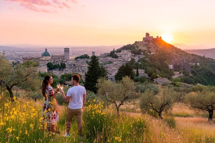 Two people stand in a grassy field, clinking wine glasses, overlooking a historic town at sunset with distant hills and a castle.