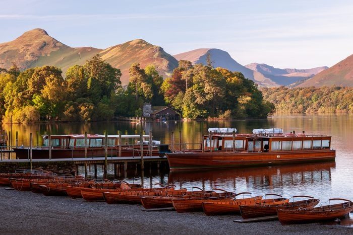 Boats rest along a calm lakeshore, surrounded by lush trees and distant mountains under a clear sky.