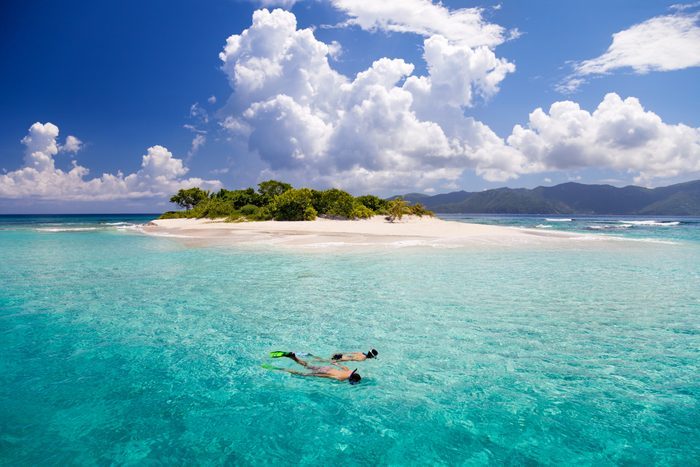 A person snorkels in clear turquoise water near a small sandy island with green vegetation under a bright sky filled with fluffy clouds.