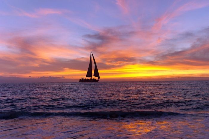 Sailboat glides on the ocean at sunset, under a vibrant sky of purple and orange hues, reflecting on gentle waves.