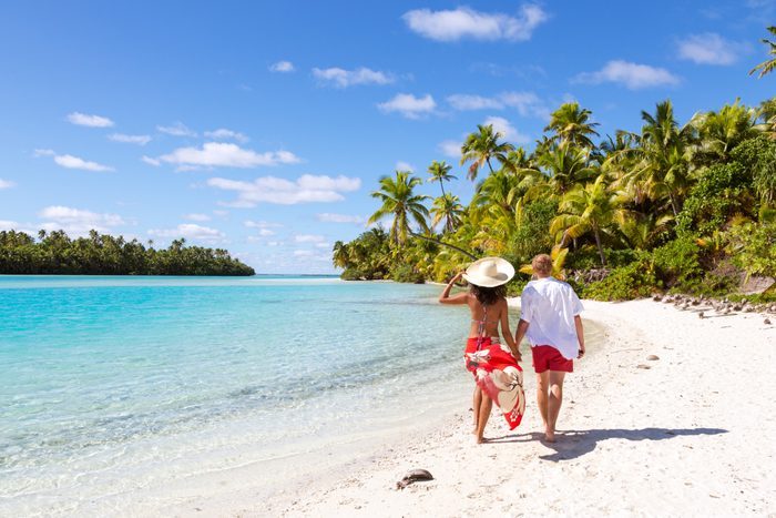 A couple walks along a sandy beach, holding hands, with turquoise water and lush palm trees under a bright blue sky.