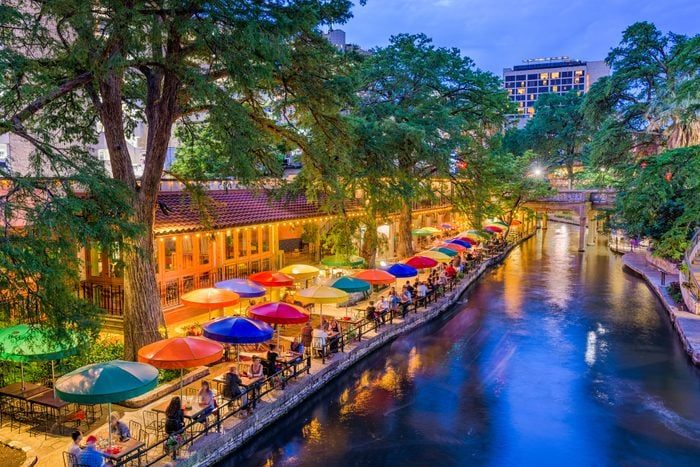Colorful umbrellas line a riverside dining area, diners sitting, amid lush trees and evening lights, with tranquil water reflecting the vibrant scene.