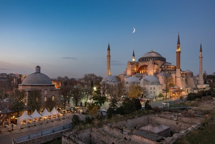 Hagia Sophia stands illuminated, surrounded by trees and historical ruins at twilight, beneath a crescent moon in a tranquil urban landscape.