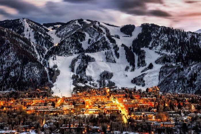 A snow-covered mountain looms over a town glowing with lights, under a dramatic, cloud-streaked sky during twilight.