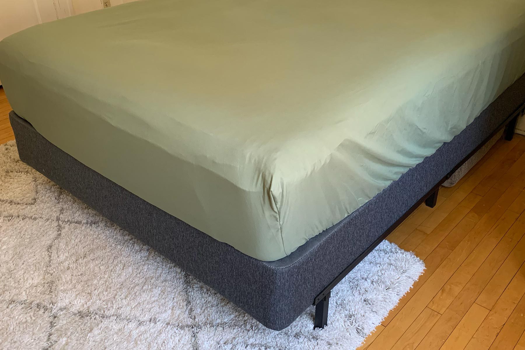 Bed with green bedsheet sits on a gray frame, positioned on a white textured carpet, in a hardwood-floored room.