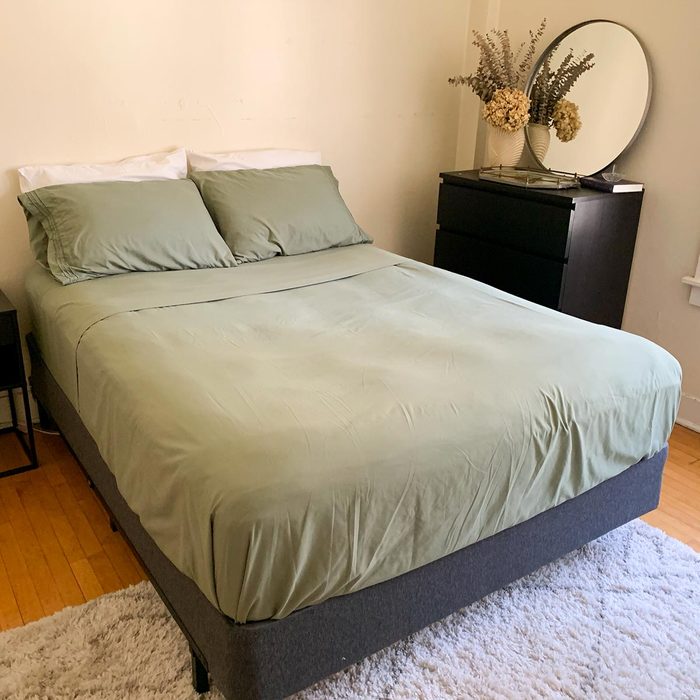 Bed is neatly made with green sheets; dresser nearby holds mirror and flowers in a minimalistic, bright bedroom.