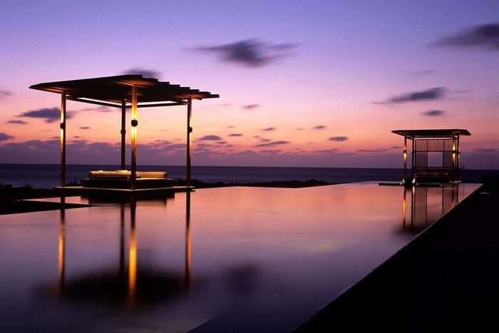 A reflecting pool under two modern pavilions glistens at sunset, with a serene ocean and colorful sky as the backdrop.