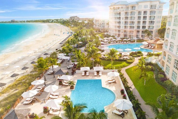 A resort features multiple pools surrounded by lounge chairs, overlooking a sandy beach and turquoise ocean under a clear, sunny sky.