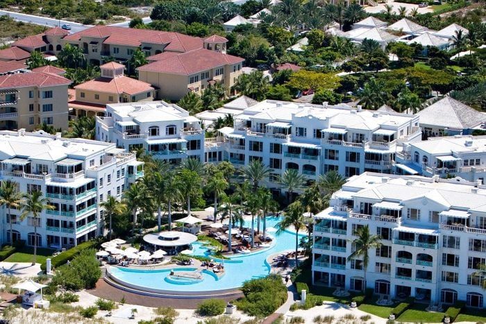 Buildings surround a curving pool area with lounge chairs and umbrellas, accented by palm trees and greenery, creating a resort-like atmosphere.
