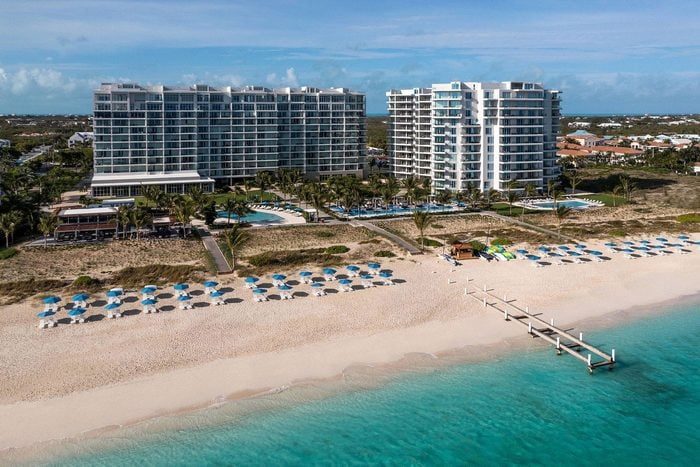 Buildings overlook a sandy beach lined with umbrellas, set beside turquoise ocean waters. The area includes pools and palm trees in a tropical setting.