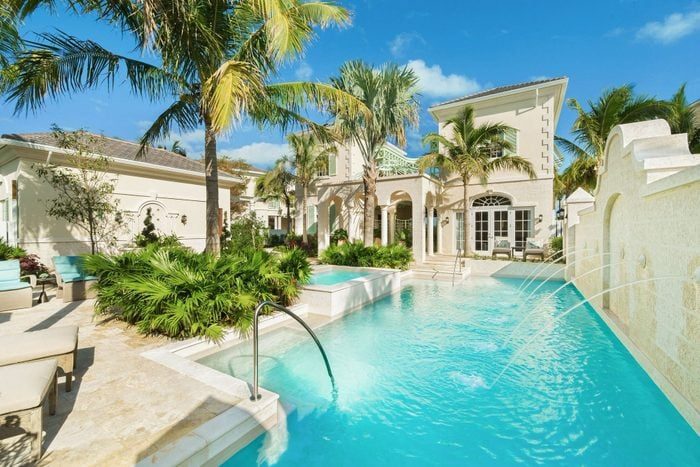 A luxurious villa features a sparkling pool surrounded by palm trees and elegant architecture, under a bright blue sky.