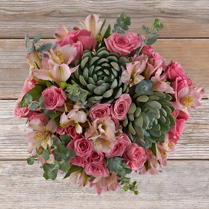 Bouquet sits composed of pink roses and succulents on rustic wooden surface, surrounded by mixed greenery, creating an elegant and natural arrangement.