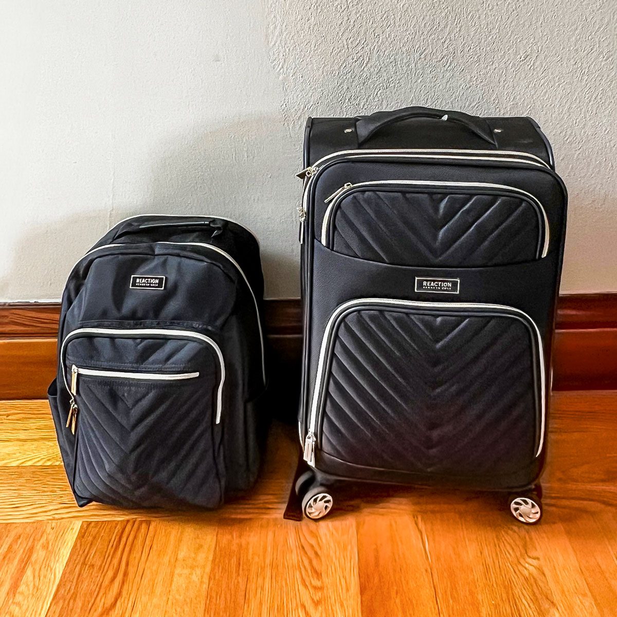 A black backpack and wheeled suitcase sit on a wooden floor against a light-colored wall. Both have quilted patterns and silver zippers.