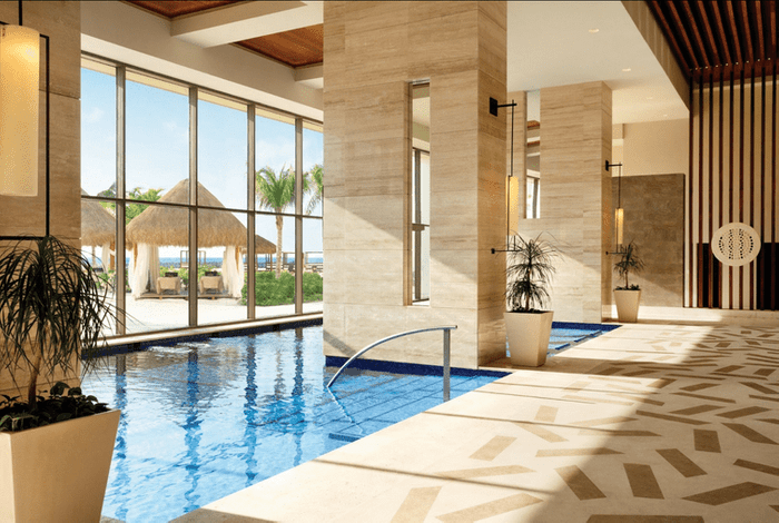Pool reflecting indoors, framed by beige columns, with large windows revealing palm trees and straw umbrellas outside, basking in sunlight.