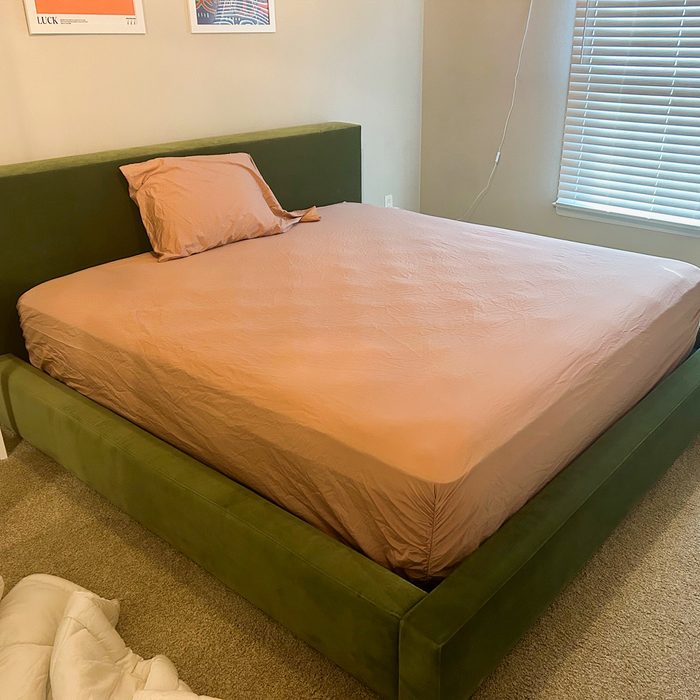 Bed with a light brown sheet and single pillow, situated in a carpeted room with framed pictures and a window with blinds.