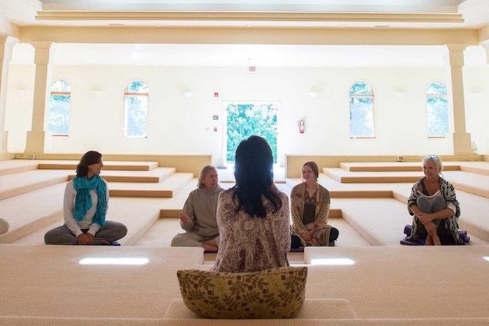 Individuals sit in a serene, spacious room with tiered wooden flooring, engaged in conversation, surrounded by large windows.