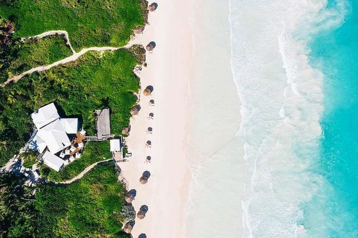 Umbrellas line the sandy beach, with waves gently lapping the shore, adjacent to a green landscape and white buildings.