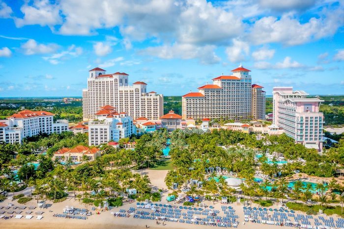 Buildings overlook pools and palm trees, with a beach in the foreground and a partly cloudy sky in the background.