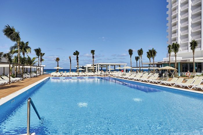 A large swimming pool sits empty, surrounded by lounge chairs and palm trees, with a tall building in the background under a clear blue sky.