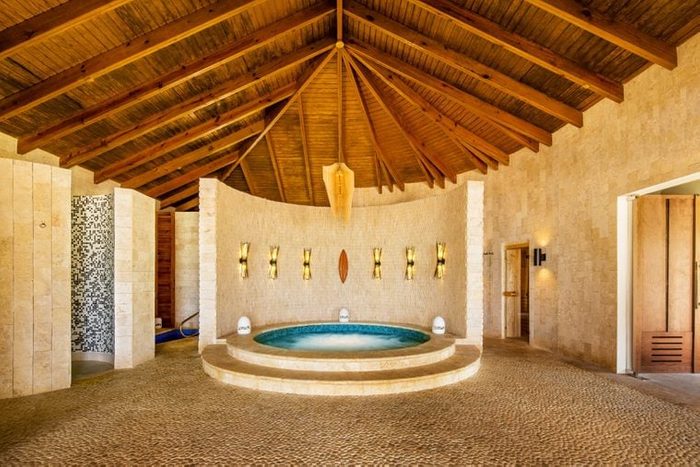 Circular jacuzzi sits inactive in rustic room with a wooden ceiling, beige stone walls, and small decorative lights. No people are present.