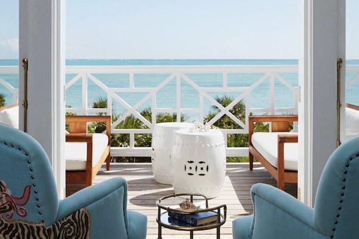 Blue chairs face a small table on a beachfront balcony, overlooking a calm ocean under a clear sky.