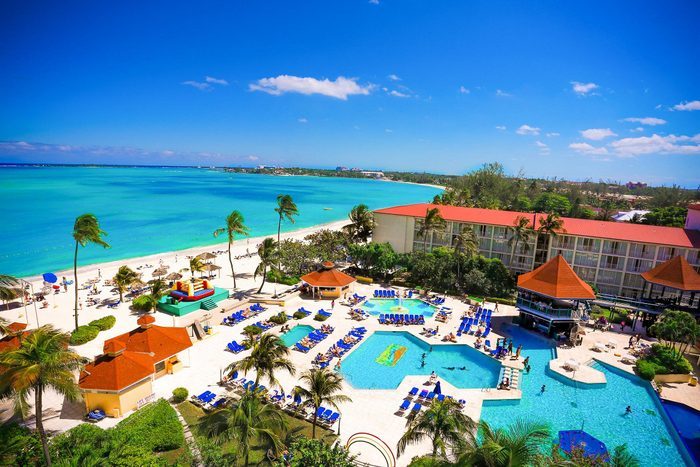 Resort pools sparkle under the sun, surrounded by palm trees, lounge chairs, and a vibrant beach, set against a clear blue sky and ocean.