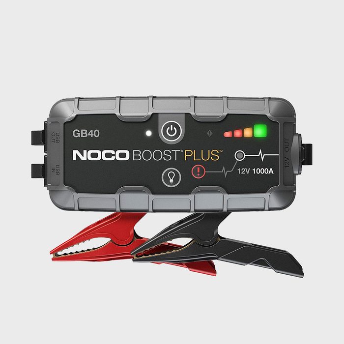 Battery jump starter, displaying lights and indicators, rests on metal clamps against a plain background. Text reads 
