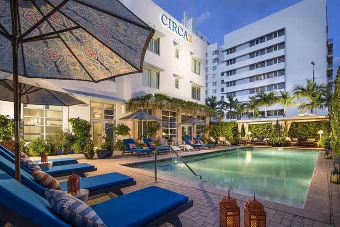 Pool reflects soft light; surrounded by blue lounge chairs under umbrellas. Building with 