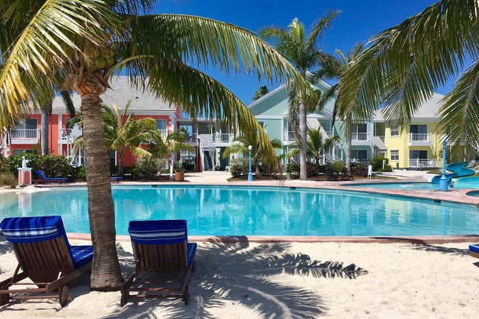 Chairs rest by a pool, surrounded by palm trees, with colorful buildings in the background under a clear blue sky.