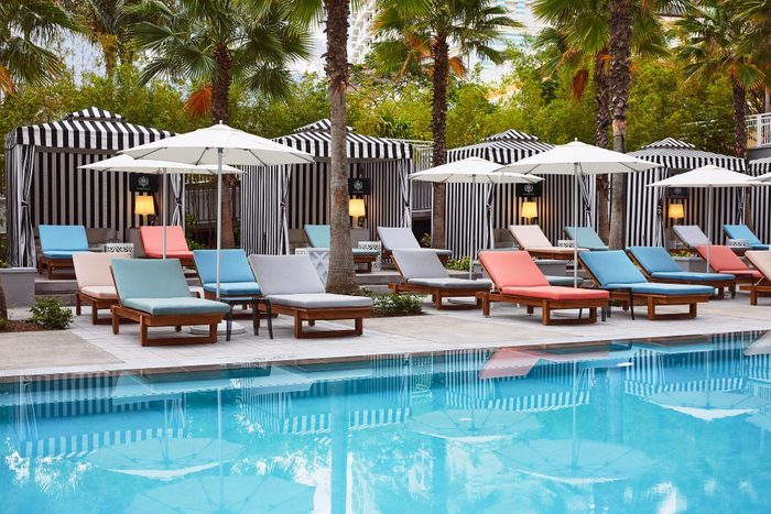 Colorful lounge chairs sit by a swimming pool with striped cabanas and palm trees in the background.