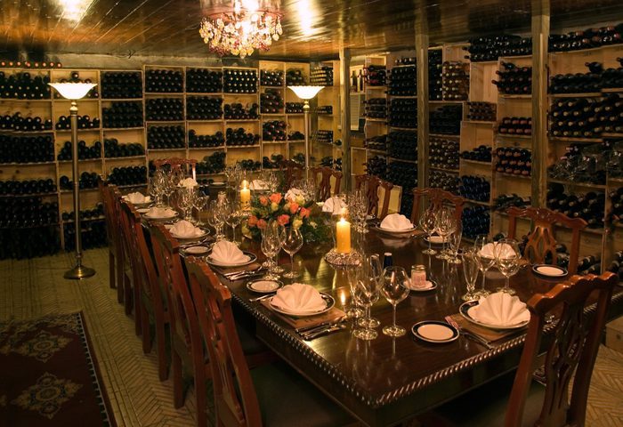 A large dining table is elegantly set with plates and glasses, surrounded by wine racks in a dimly lit, cozy wine cellar.