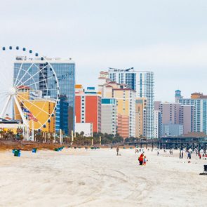 Ferris wheel stands prominently beside colorful high-rise buildings along a busy beach with people walking and relaxing.