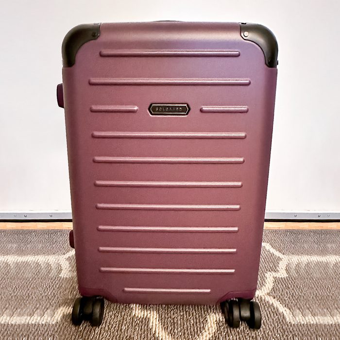 Suitcase standing upright, featuring rolling wheels, maroon color, in front of a plain white wall on a patterned rug. Text reads 