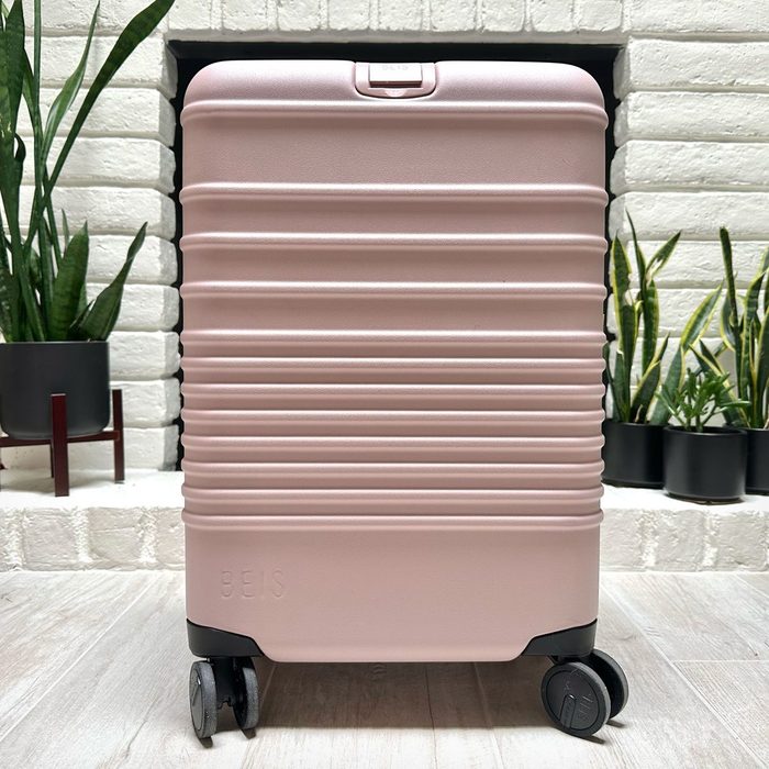 A pink suitcase stands on wheels against a white brick wall, surrounded by potted plants.