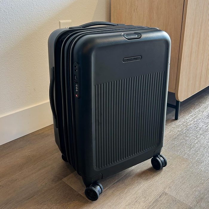 Black suitcase standing on four wheels, resting on a wooden floor, beside a light-colored wall and cabinet.