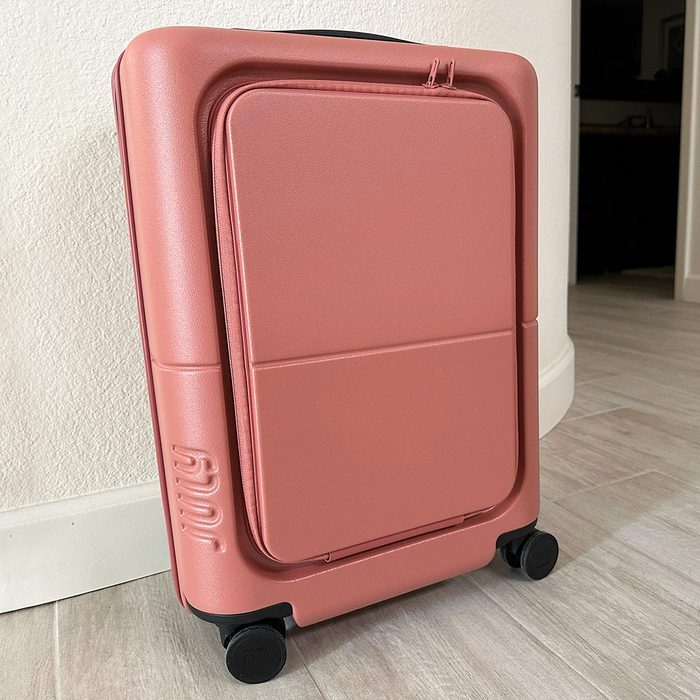 Suitcase stands on floor; it has a sleek, pink design with four wheels and the brand 