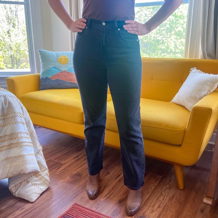 Person stands with hands on hips, wearing dark jeans and boots, in front of a yellow couch in a sunny, modern living room.