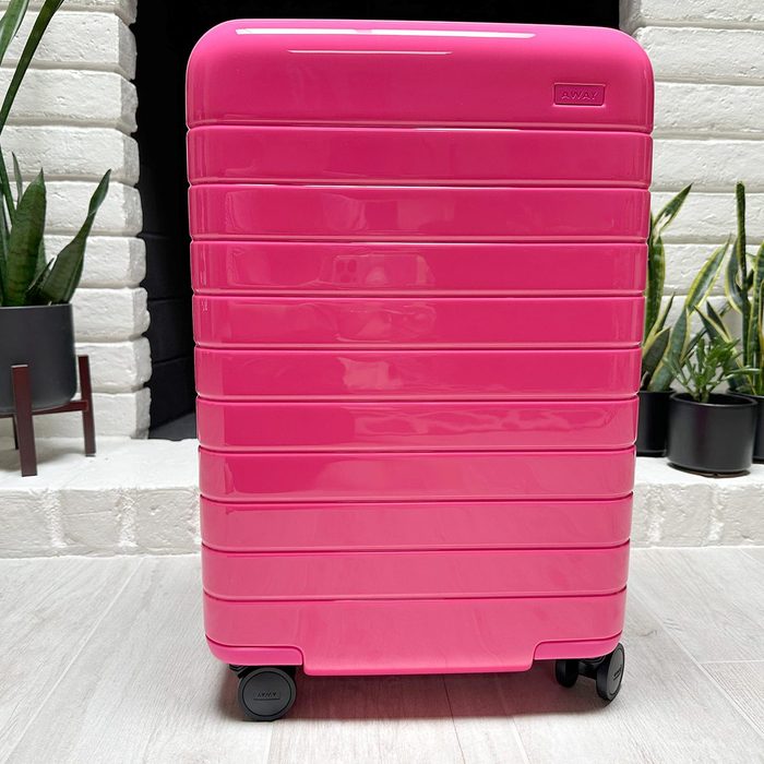 Pink suitcase stands on the floor, surrounded by white brick and potted plants; features four wheels. Text: 