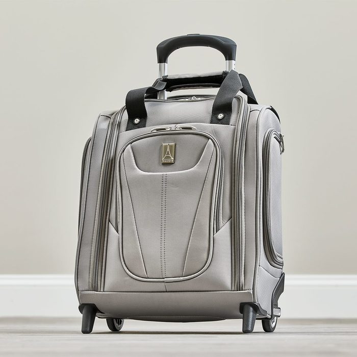 A gray suitcase stands upright with wheels, an extended handle, and front zippered pockets, positioned in a simple, beige room setting.