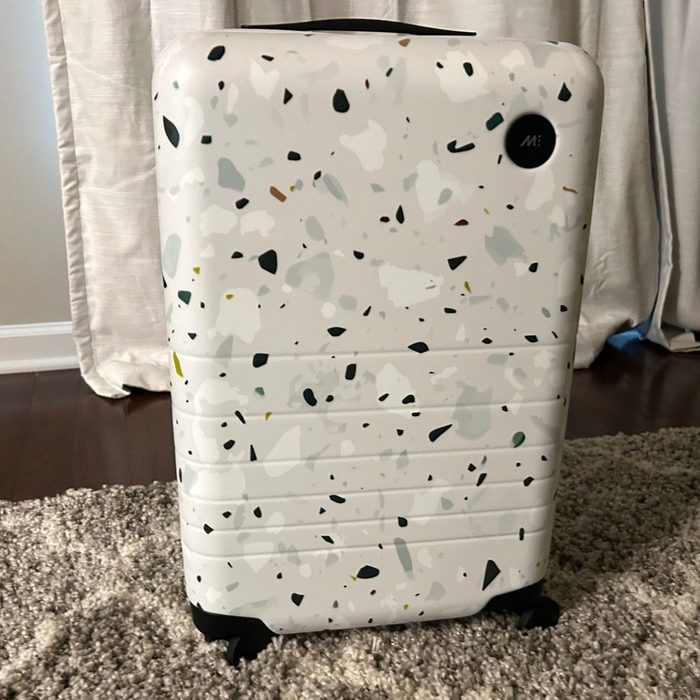 A speckled suitcase stands upright on a carpeted floor, near beige curtains in a room.