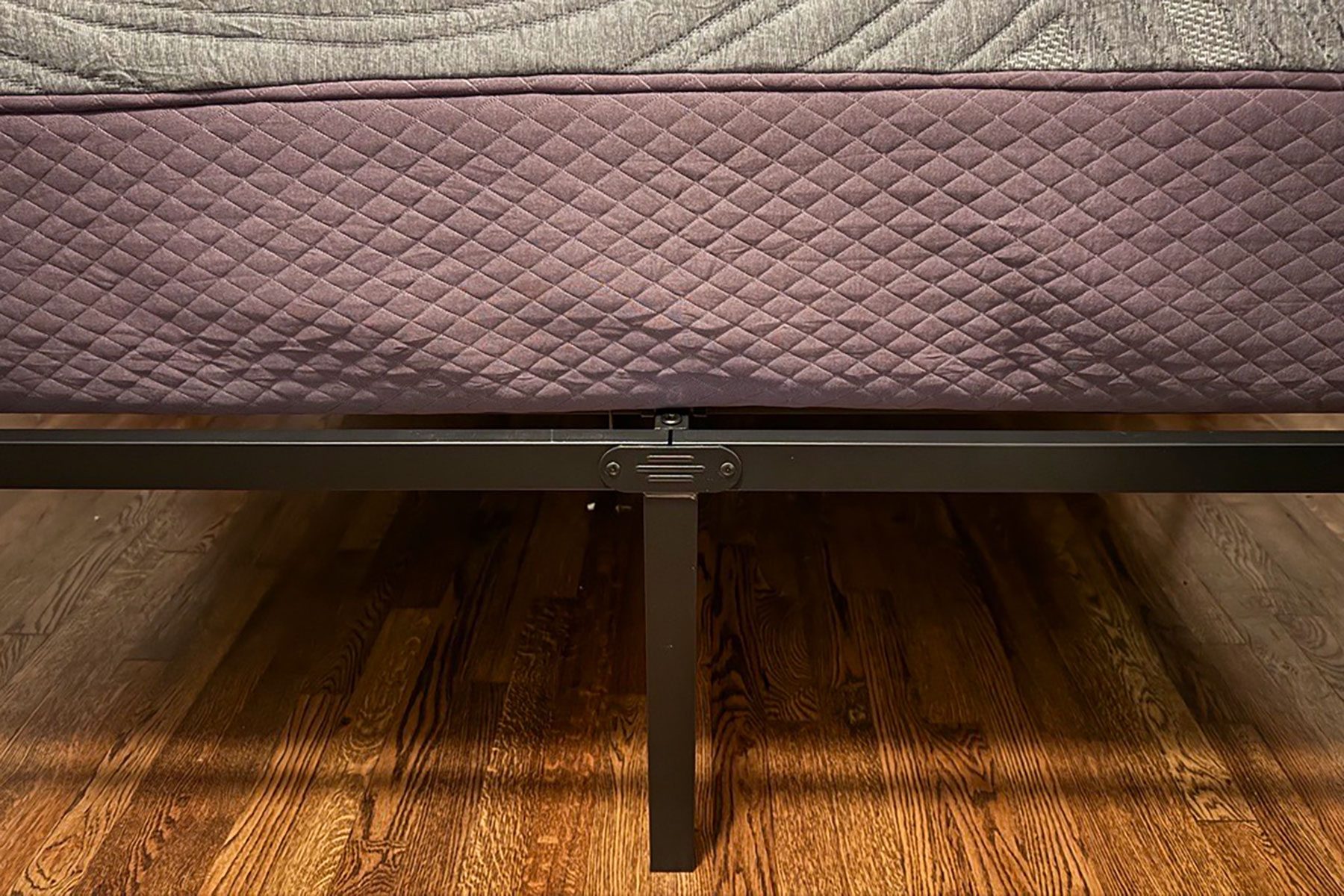 Bed frame supports a quilted fabric base, standing on wooden flooring in a dimly lit room.
