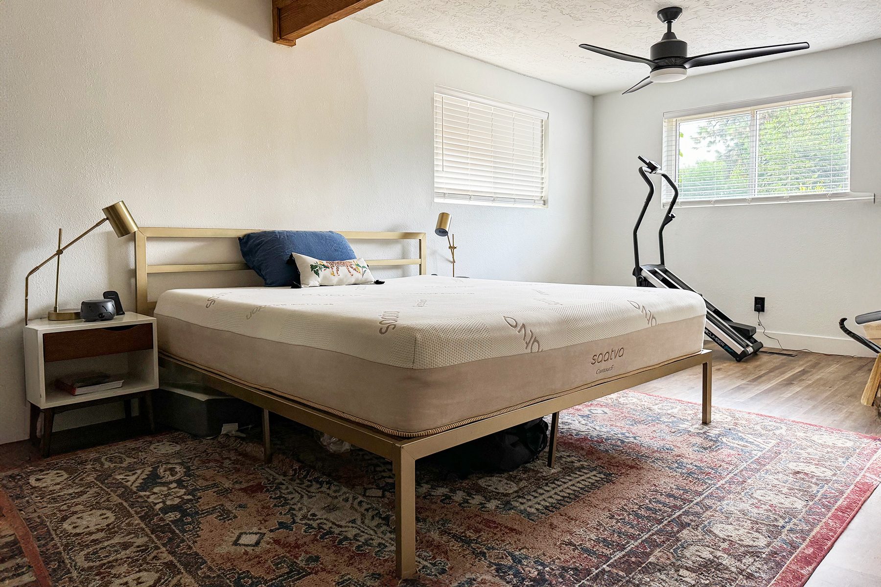 Bed stands on ornate rug; room features exercise equipment, nightstands with lamps, and large windows.