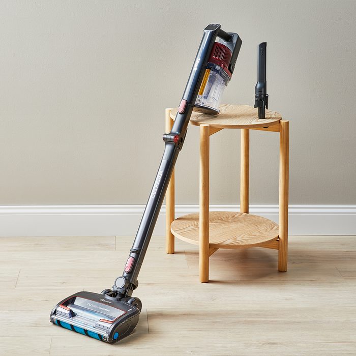A cordless vacuum cleaner leans against a small wooden table in a minimalist room with a light-colored wall and floor.