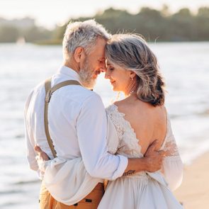 A couple embraces tenderly by the water at sunrise, wearing elegant clothing, with gentle waves and trees in the background.
