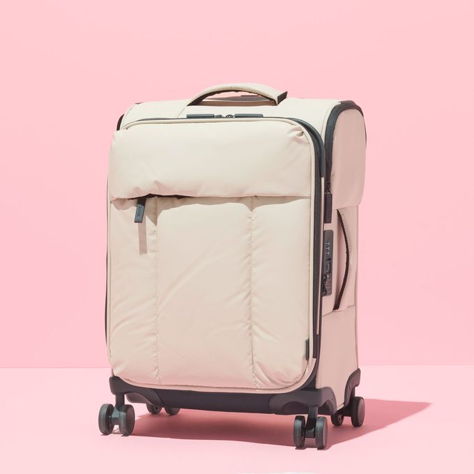A beige suitcase stands on four wheels against a pink background, featuring a zipper and handle on top.