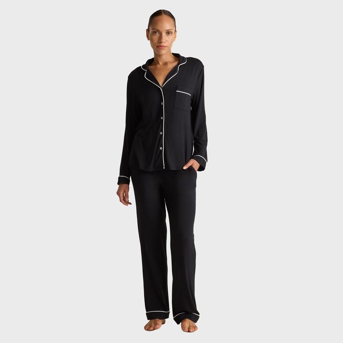 A person wears black pajama set with white piping, standing barefoot against a plain white background.