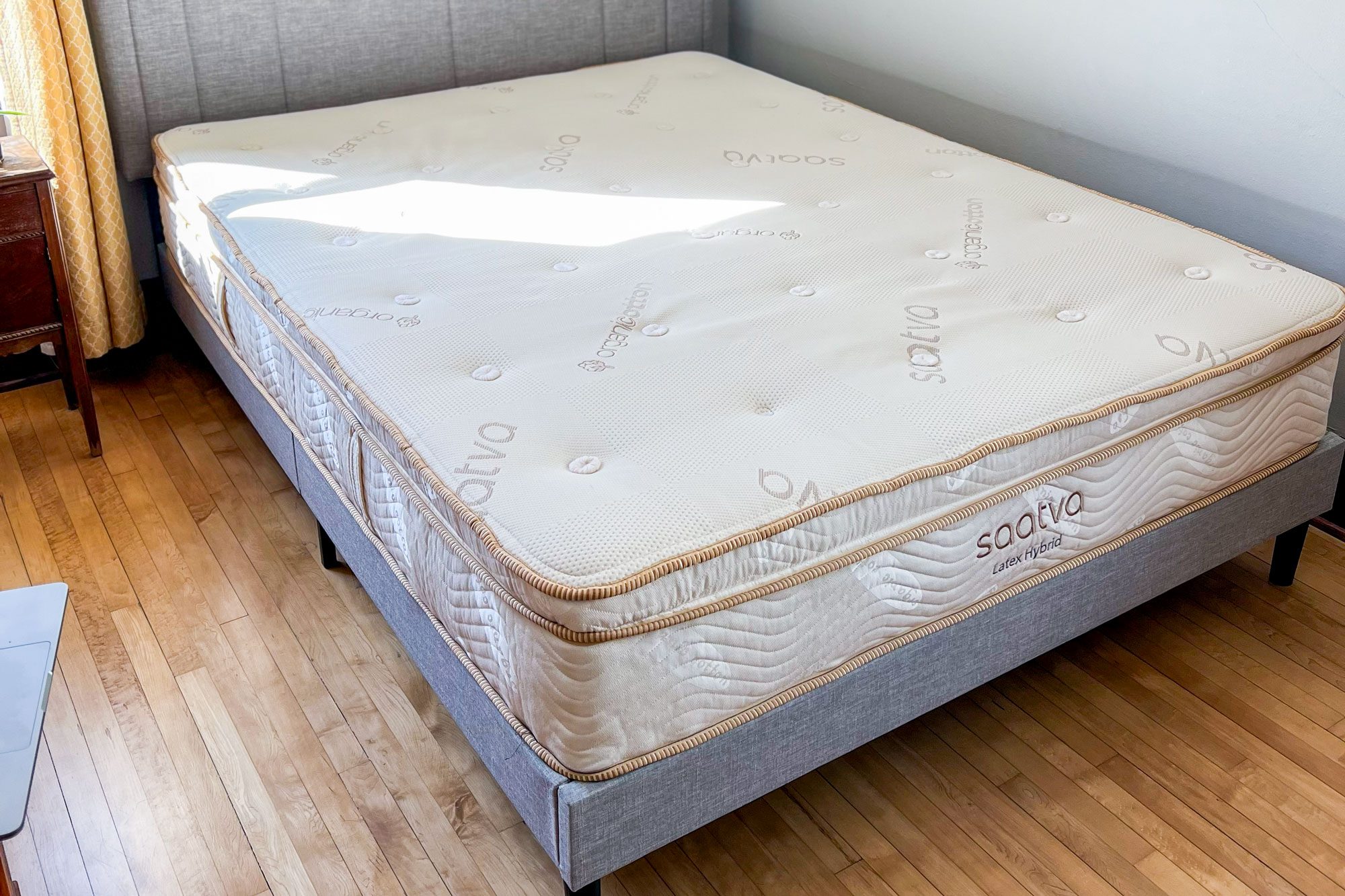 Mattress rests on a grey bed frame in a sunlit bedroom, with wooden floor and a yellow curtain nearby. Text: "Saatva Latex Hybrid."